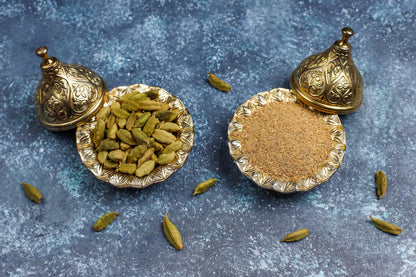 Decorative containers with green cardamom pods and brown powder on a textured surface